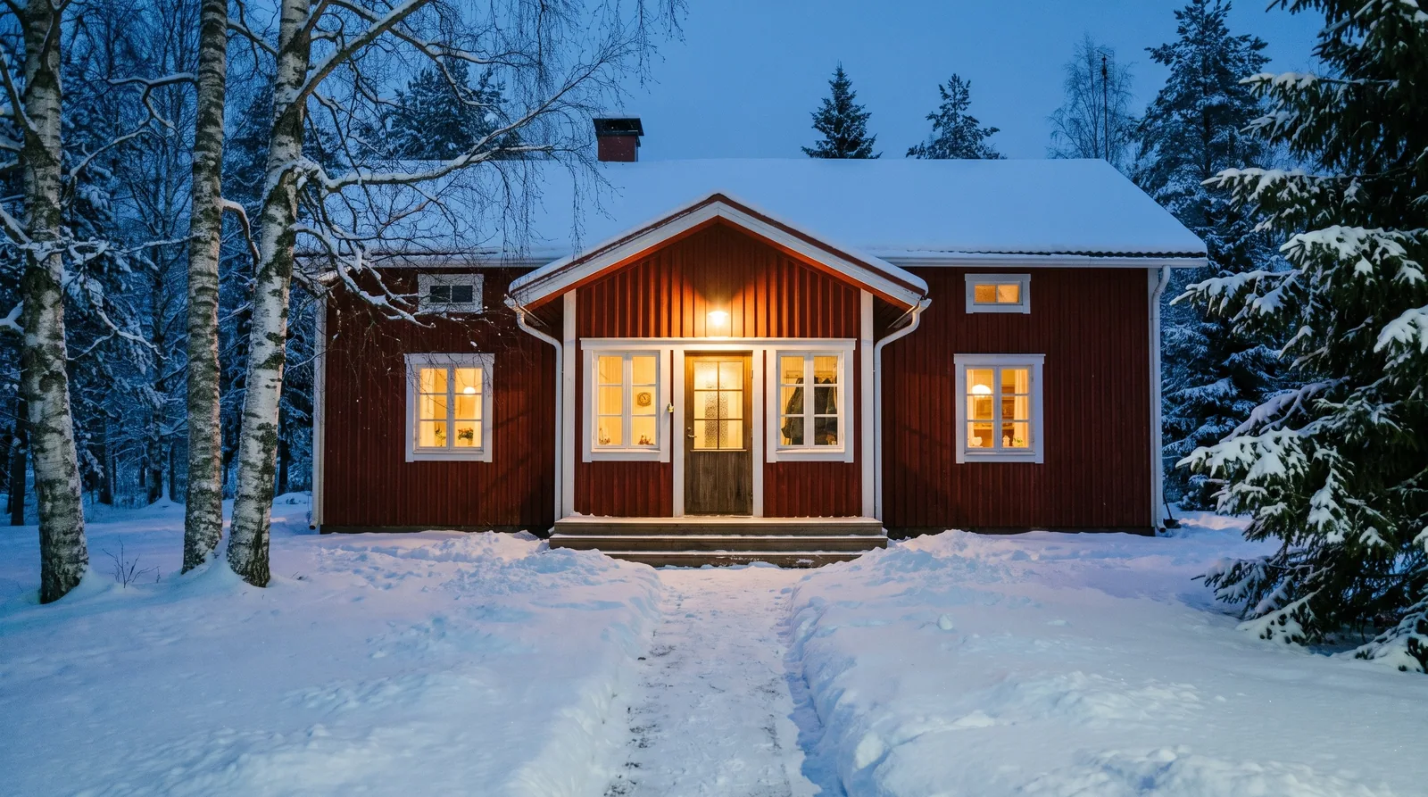 Punainen puutalo lumisessa pihapiirissä tammikuun sinisellä hetkellä, ikkunoissa lämmin valo