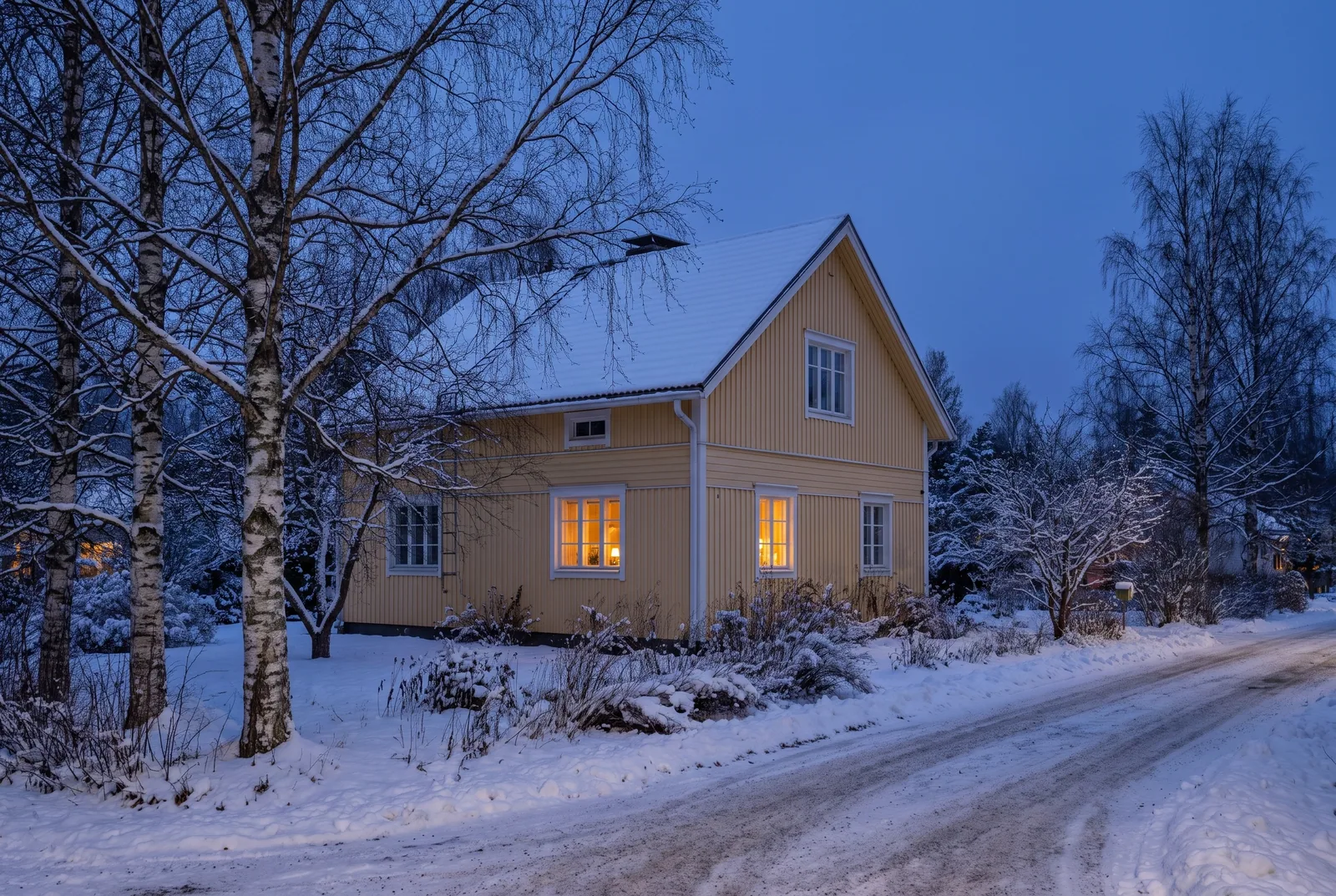 Vaaleankeltainen rintamamiestalo lumisessa pihassa talvihämärässä, ikkunoissa lämmin valo